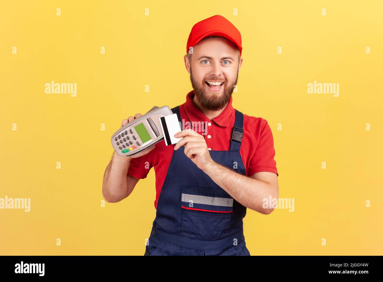 Excited joyful bearded worker man holding pos terminal for contactless payment and credit card, paying for service, expressing happiness. Indoor studio shot isolated on yellow background. Stock Photo