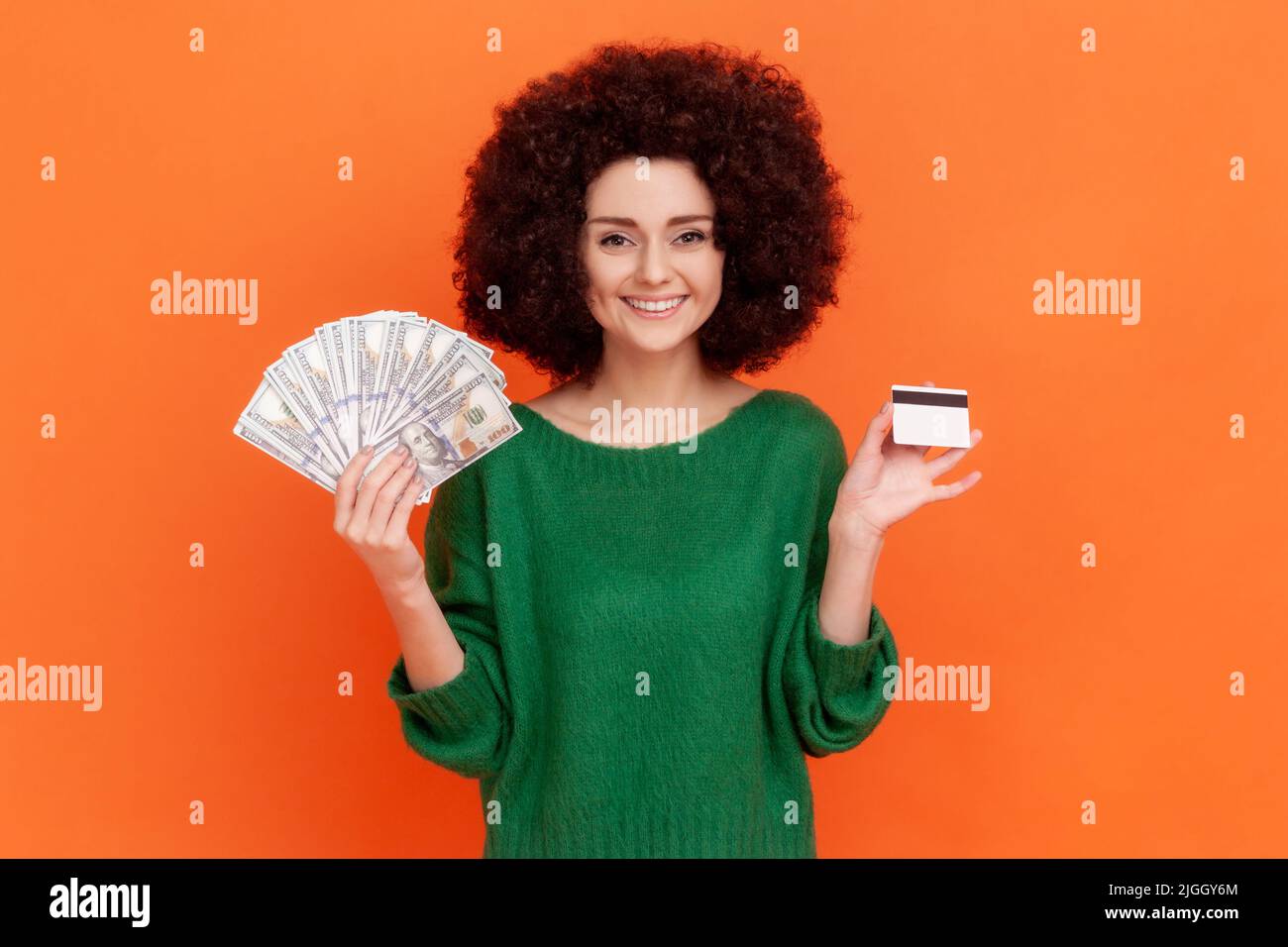 Satisfied woman with Afro hairstyle wearing green casual style sweater showing to camera dollar banknotes and credit card, investments. Indoor studio shot isolated on orange background. Stock Photo