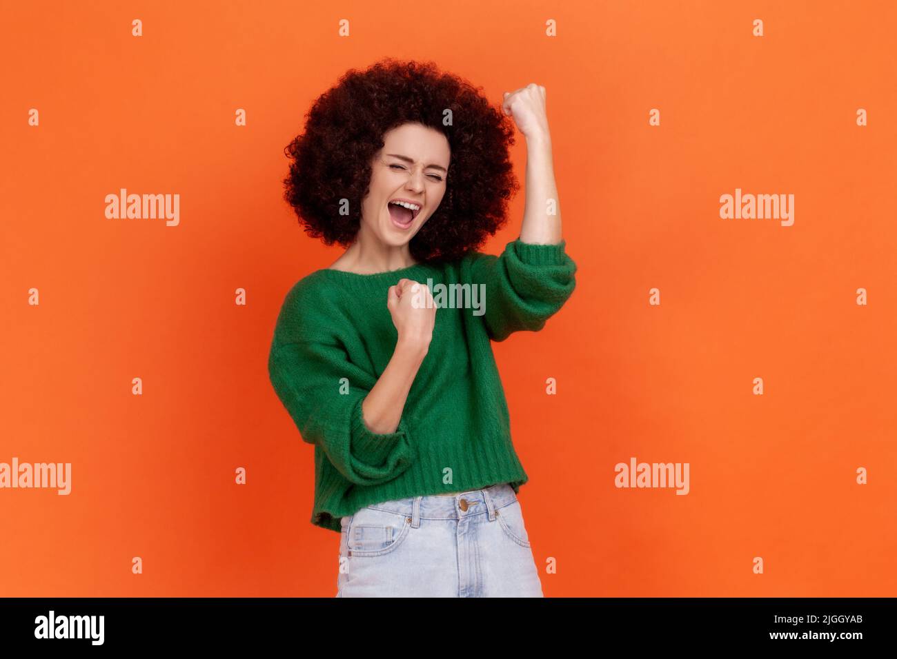 Portrait of happy excited woman with Afro hairstyle wearing green casual style sweater celebrating her winning, clenched fists, yelling. Indoor studio shot isolated on orange background. Stock Photo
