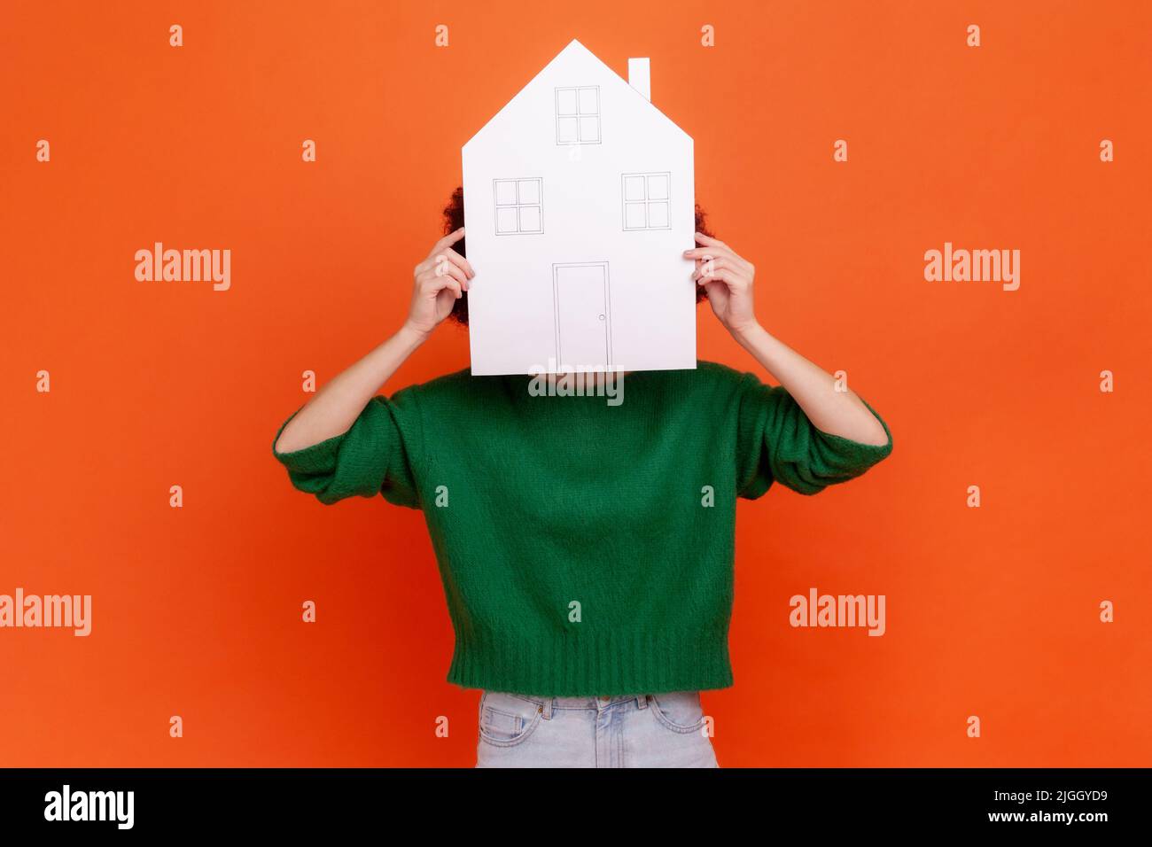Anonymous woman wearing green casual style sweater hiding her face behind paper house, buying new flat, property, real estate agency. Indoor studio shot isolated on orange background. Stock Photo