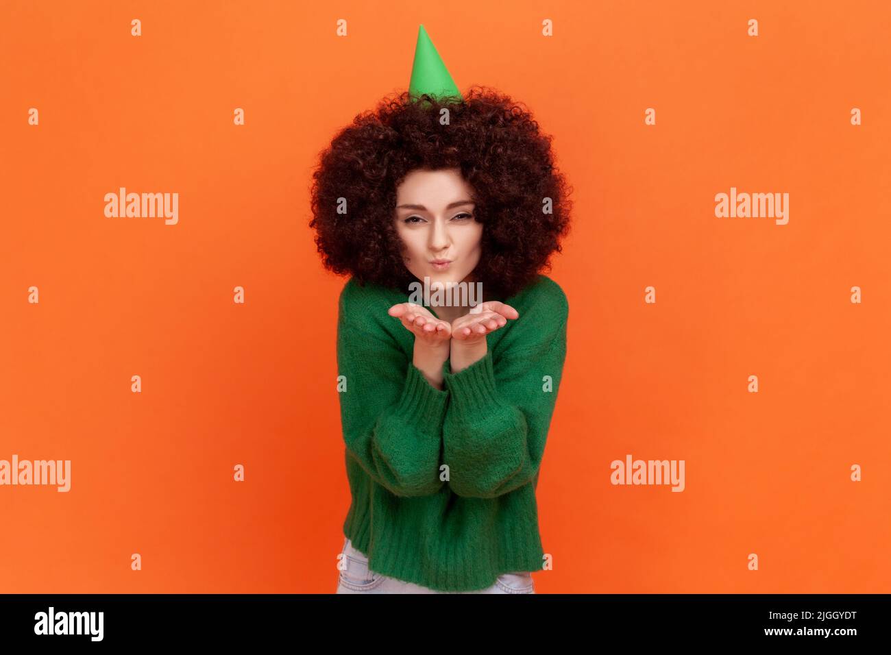 Happy birthday woman with Afro hairstyle wearing green casual style sweater thanks for the congratulations, sending air kisses to her guests. Indoor studio shot isolated on orange background. Stock Photo