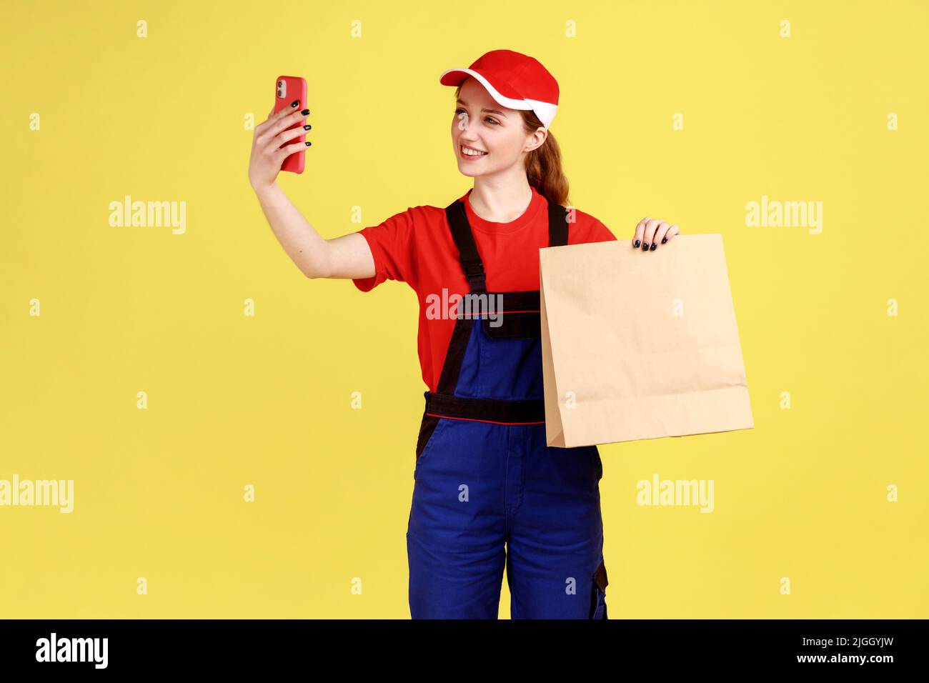 Portrait of delivery woman posing with paper package in hands, using mobile phone for communication via video call, wearing overalls and red cap. Indoor studio shot isolated on yellow background. Stock Photo