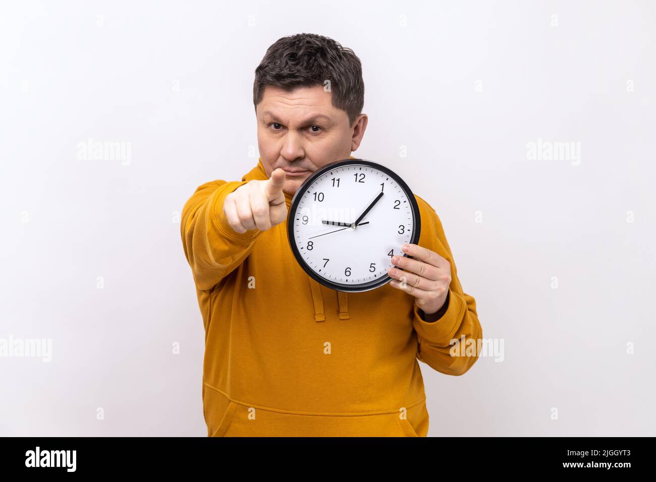 Portrait of happy positive man holding big wall clock and pointing to camera with pleasant smile, time to go, wearing urban style hoodie. Indoor studio shot isolated on white background. Stock Photo