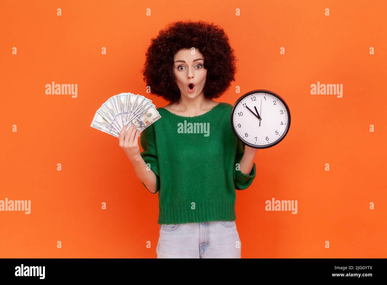 Shocked astonished woman with Afro hairstyle wearing green casual style sweater holding wall clock and dollar banknotes, surprised expression. Indoor studio shot isolated on orange background. Stock Photo