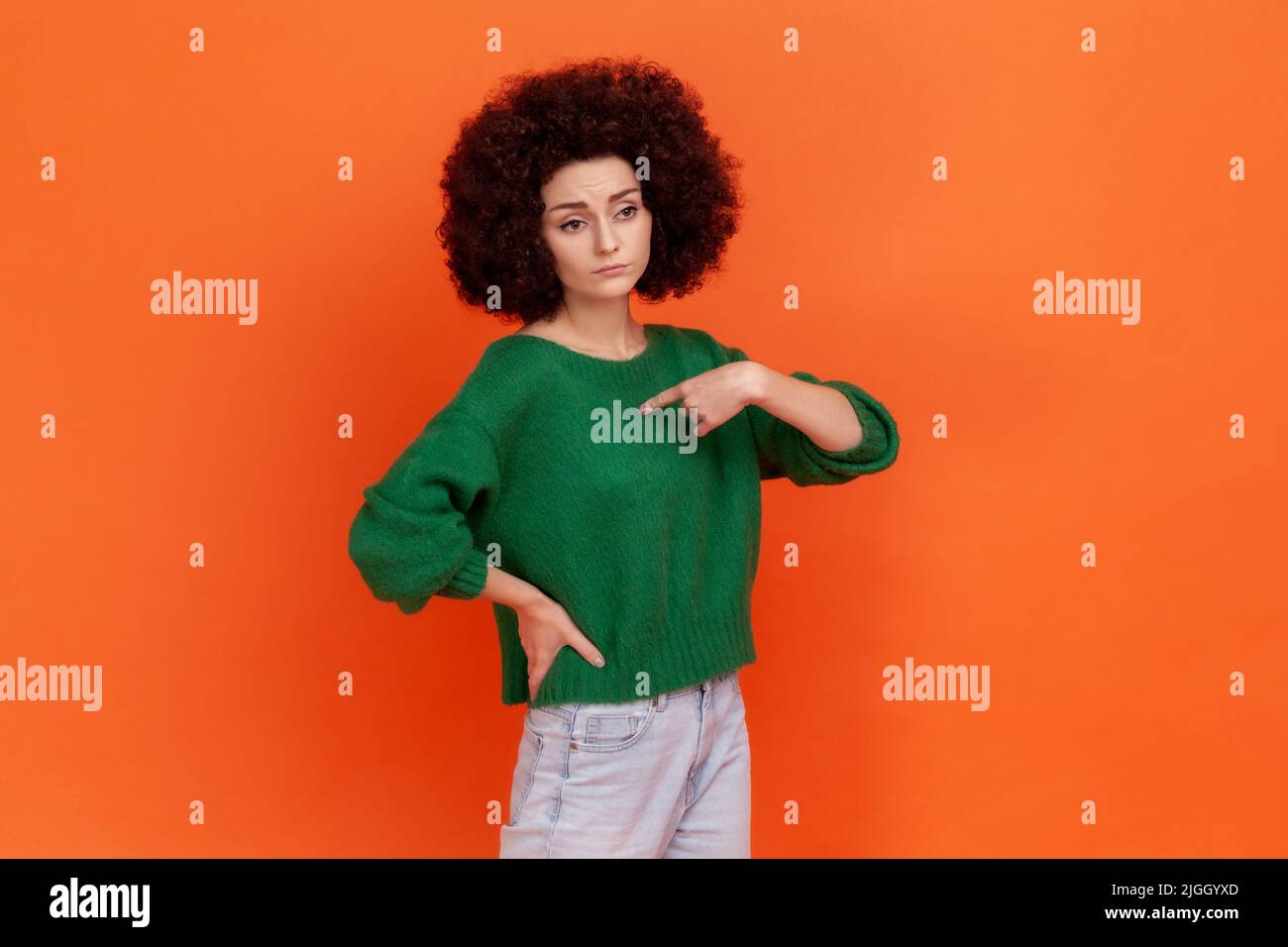Portrait of selfish woman with Afro hairstyle wearing green casual style sweater pointing at herself with finger, having proud expression. Indoor studio shot isolated on orange background. Stock Photo