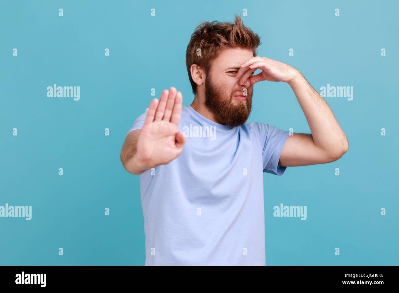 Portrait of handsome bearded man standing, pinching nose and rejecting or banning, showing stop ban gesture with palm, disgusting expression. Indoor studio shot isolated on blue background. Stock Photo