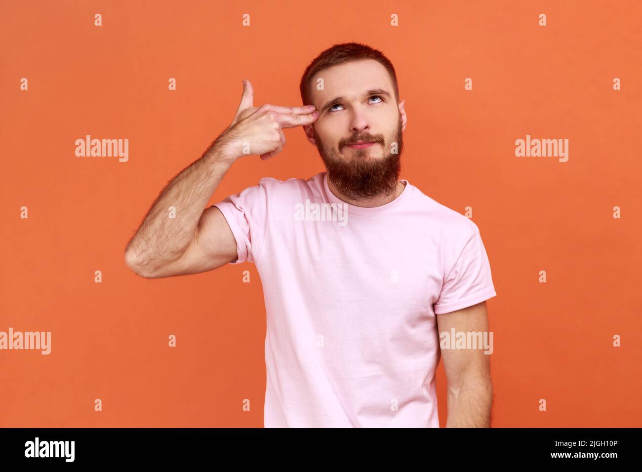 Suicide gesture. Portrait of frustrated desperate bearded man pointing finger gun to head, feeling depressed and shooting himself, wearing T-shirt. Indoor studio shot isolated on orange background. Stock Photo