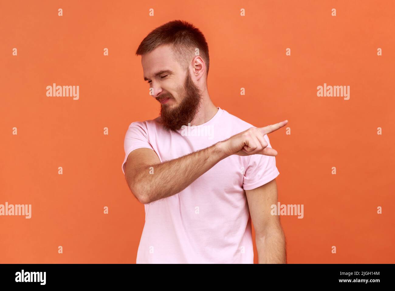 Portrait of bearded man pointing finger aside, ordering get out and looking resentful, boss dismissing from work, showing exit, wearing pink T-shirt. Indoor studio shot isolated on orange background. Stock Photo