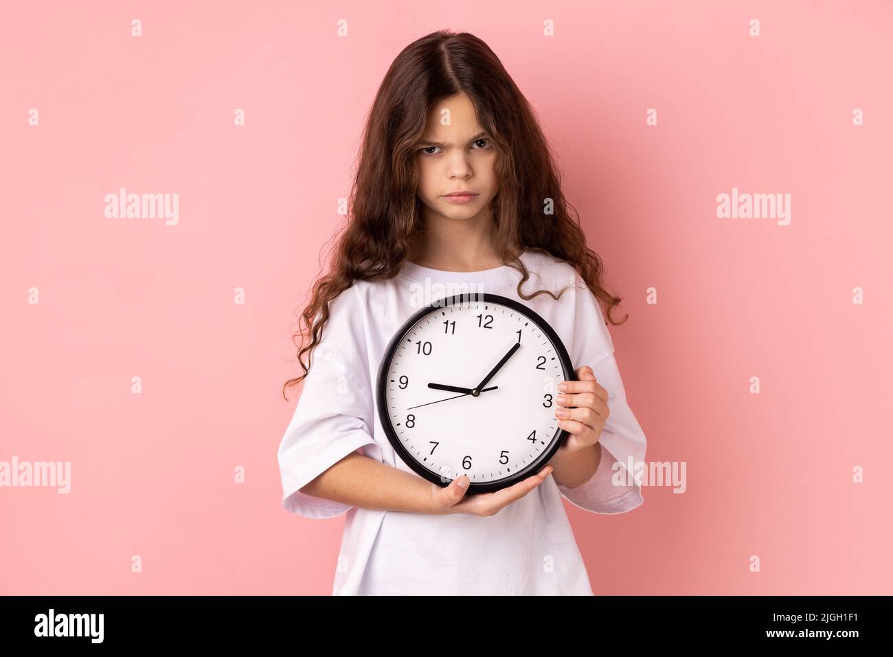 Portrait of little girl wearing white T-shirt holding wall clock, being unhappy, deadline, being sad, not finished her task in time. Indoor studio shot isolated on pink background. Stock Photo