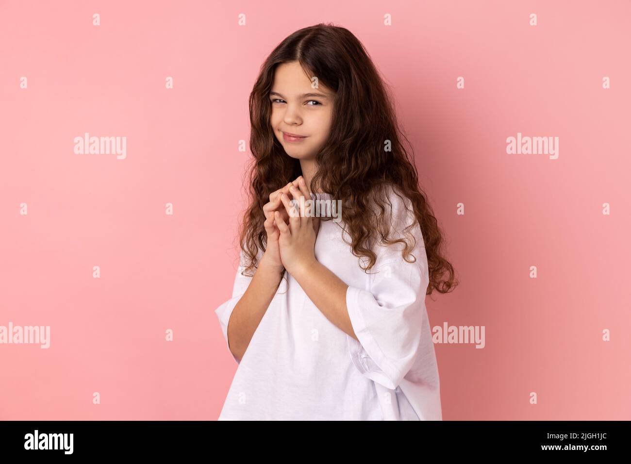 Portrait of cunning little girl in T- shirt having tricky plans, looking at camera with cheating face, smirking mysteriously, scheming cheats. Indoor studio shot isolated on pink background. Stock Photo