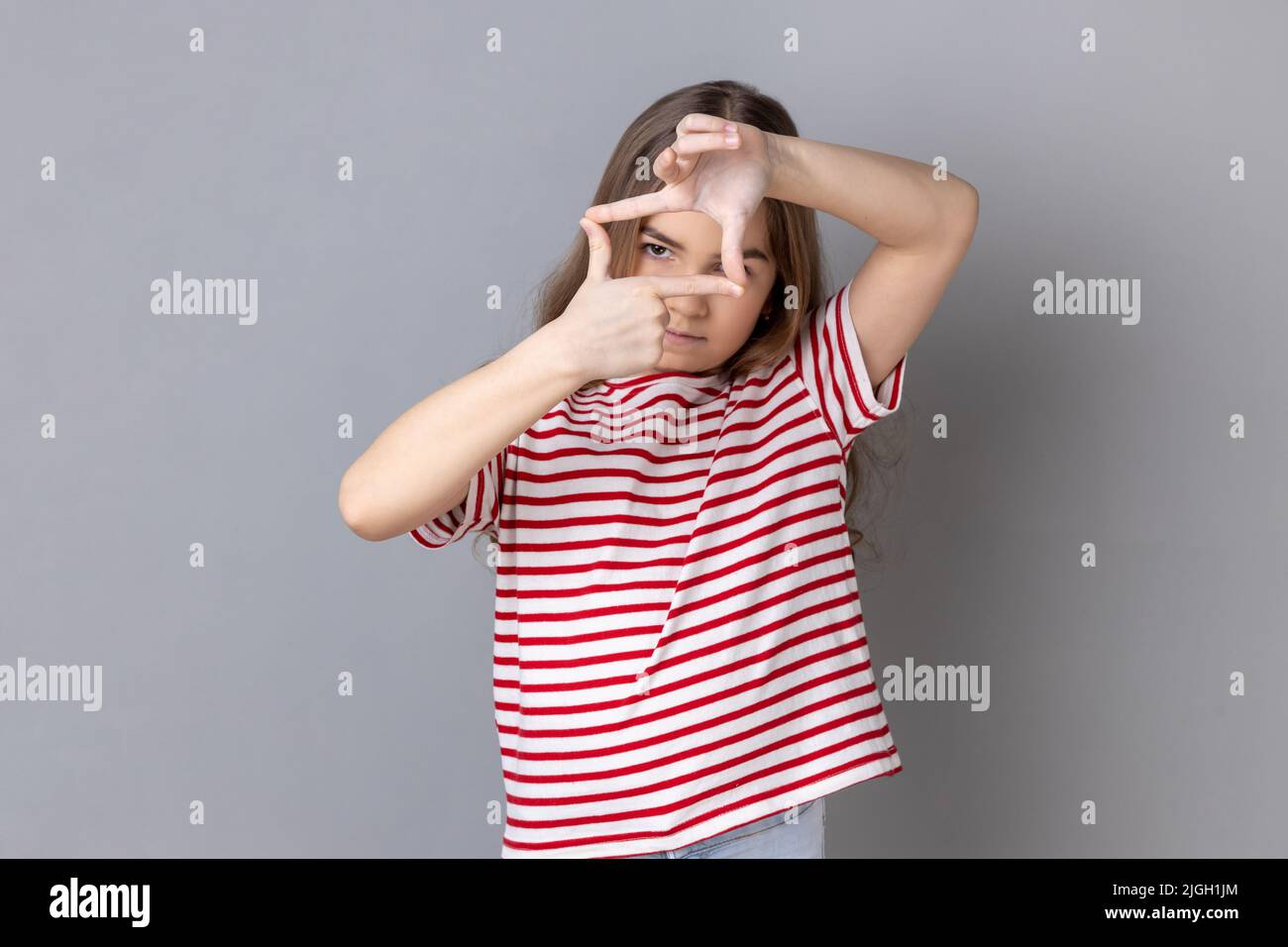 Portrait of little girl wearing striped T-shirt looking through fingers imagining frame to make good photos, dreaming to be a photographer. Indoor studio shot isolated on gray background. Stock Photo