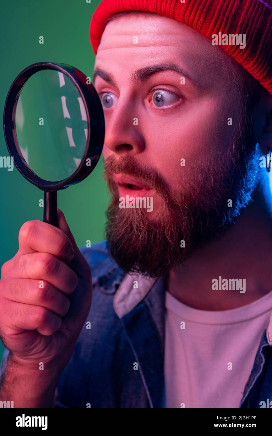 Side view of hipster man standing, holding magnifying glass and looking aside and having astonished shocked face. Indoor studio shot isolated on colorful neon light background. Stock Photo