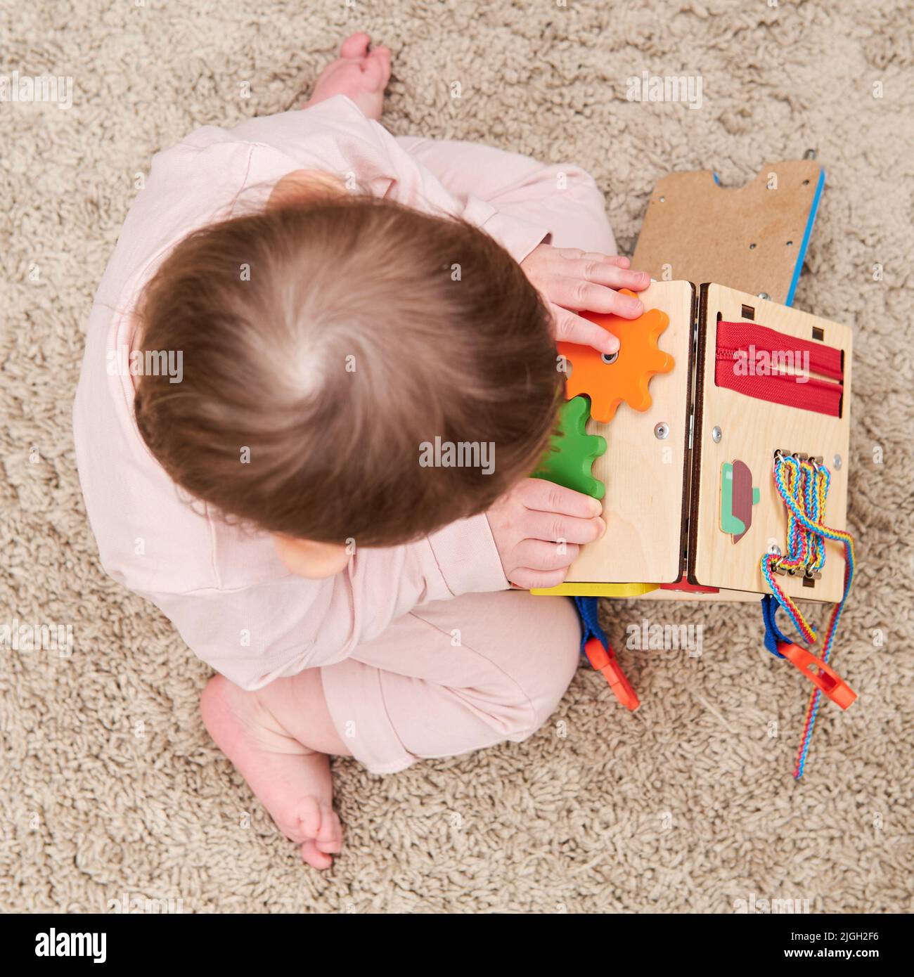 Happy toddler baby boy is playing with a busyboard cube on the nursery floor. Educational toys for children, wooden game panel. A child aged one year Stock Photo