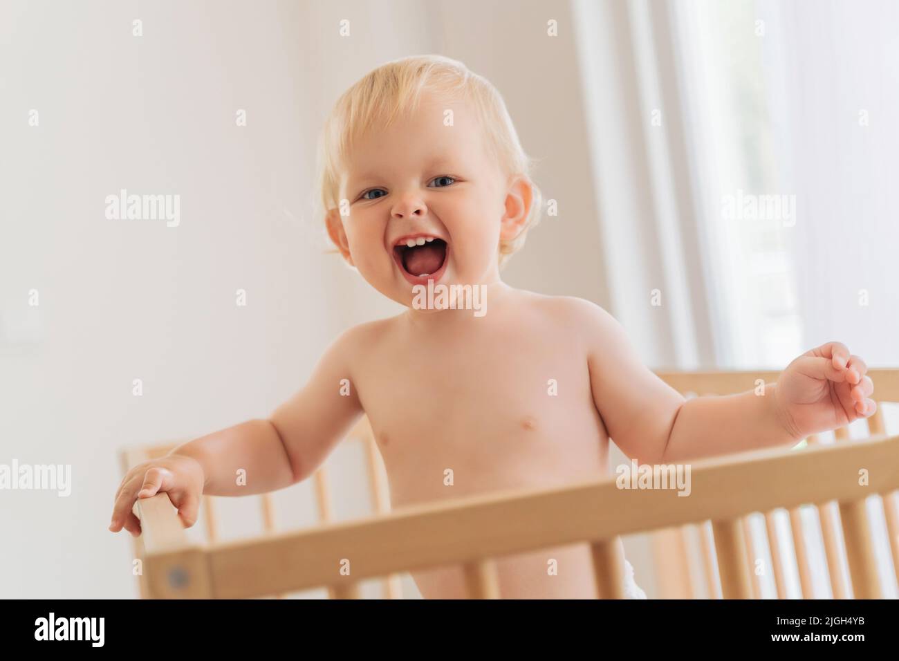 Portrait of funny 11 month old blond baby standing in bed holding onto bumpers shouting with excitement happy to see his father returned home from work. Carefree happy childhood Stock Photo