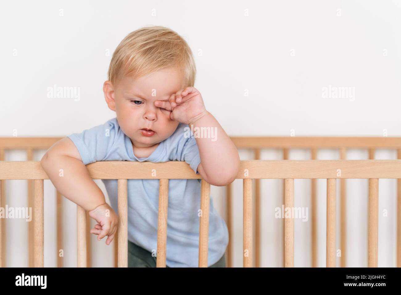 Portrait of beautiful cute little 11 months old sleepy tired baby boy in blue clothes rubbing eyes falling asleep standing in bed leaning on bumpers isolated on white background. Time to nap Stock Photo