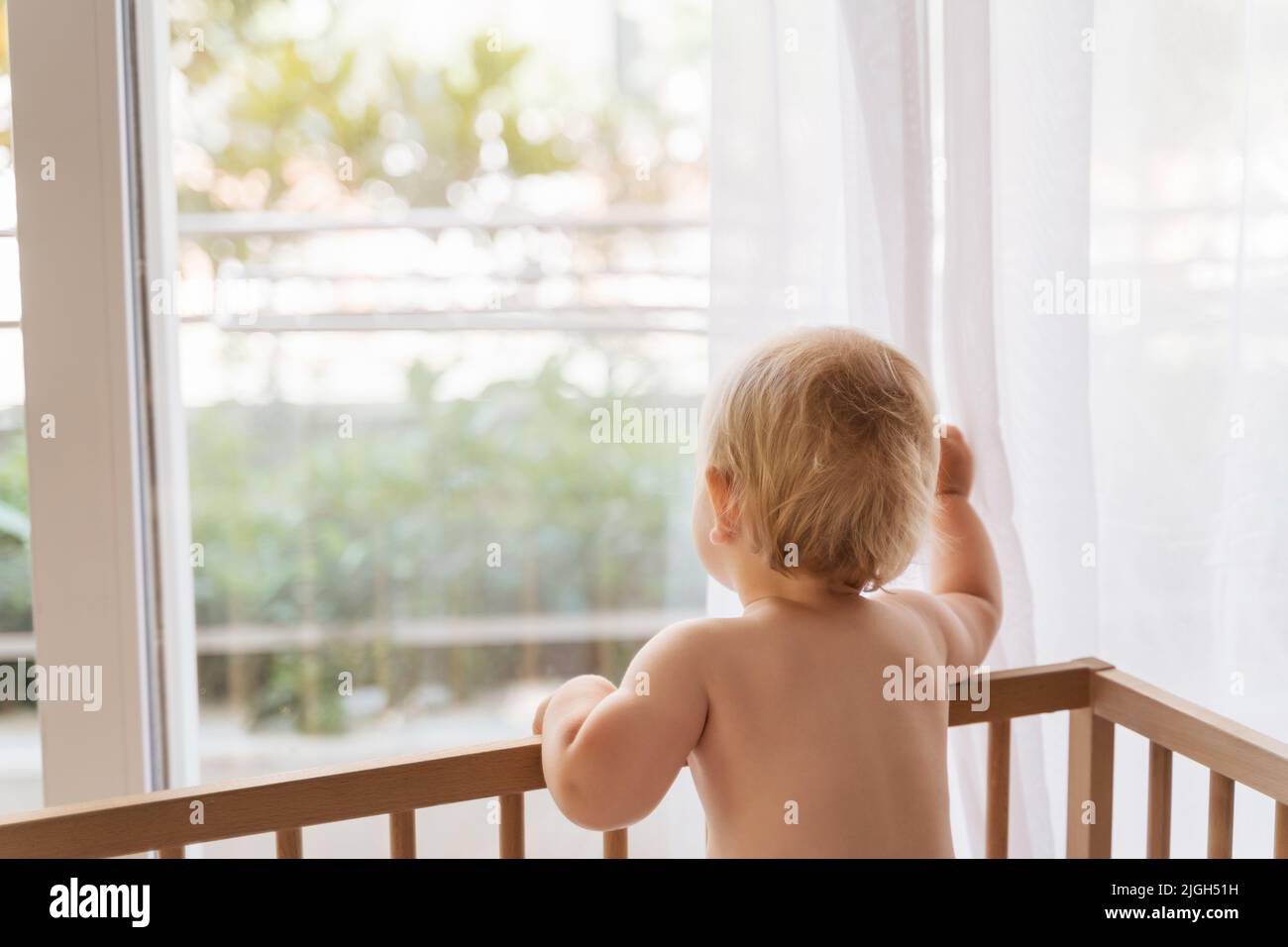 Back view of sad lonely baby standing in bed looking through window