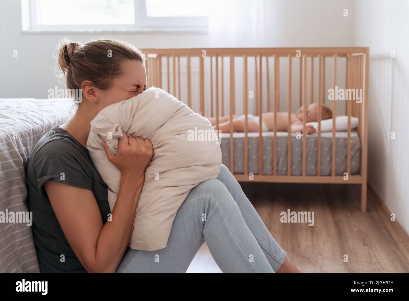 Depressed mother screaming with anger and desperation in pillow to overcome negative emotions, suffering postnatal depression because of lack of help and support with childcare routine Stock Photo