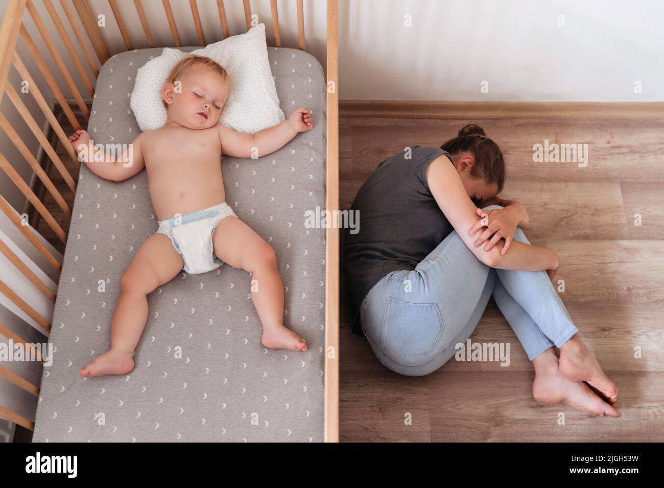 Upper view of cute baby napping in his bed next to his mother lying on floor crying, feeling desperate and lonely, suffering postnatal depression. Difficulties of maternity Stock Photo
