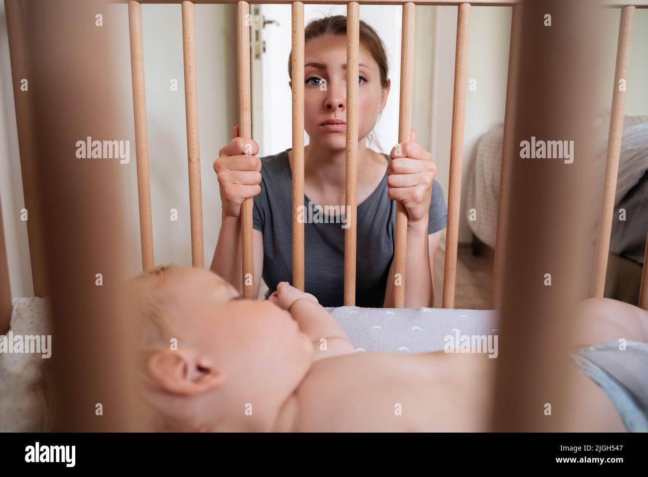 Young frustrated mother feels imprison while takes care of her infant and suffering from postnatal depression. Depressed woman looking at camera while her little toddler baby sleeping in his bed Stock Photo