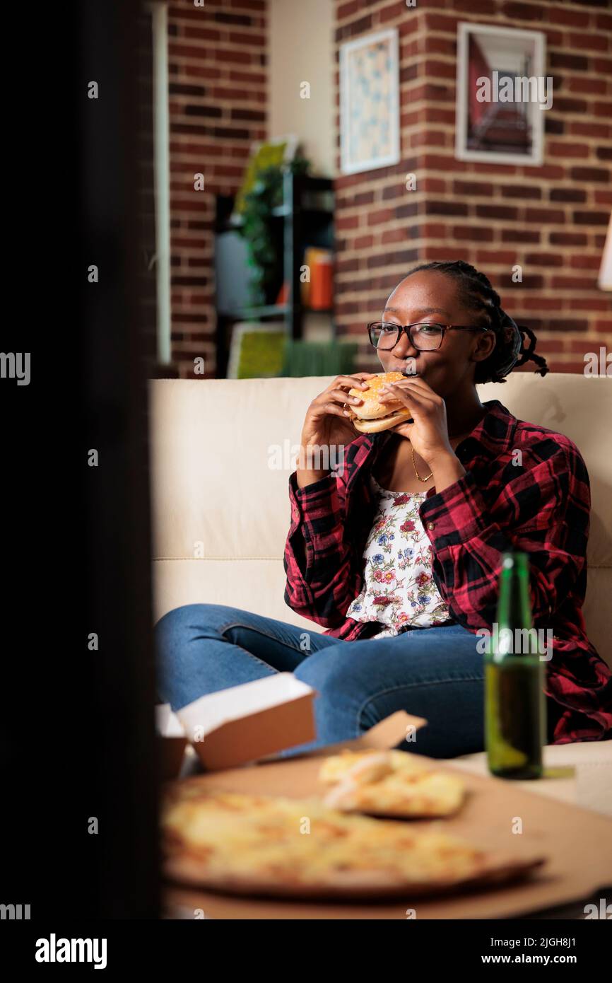 Happy young adult eating burger and drinking bottle of beer, enjoying leisure activity and fun movie on television. Having takeaway fast food delivery meal with beverage and snacks at home. Stock Photo