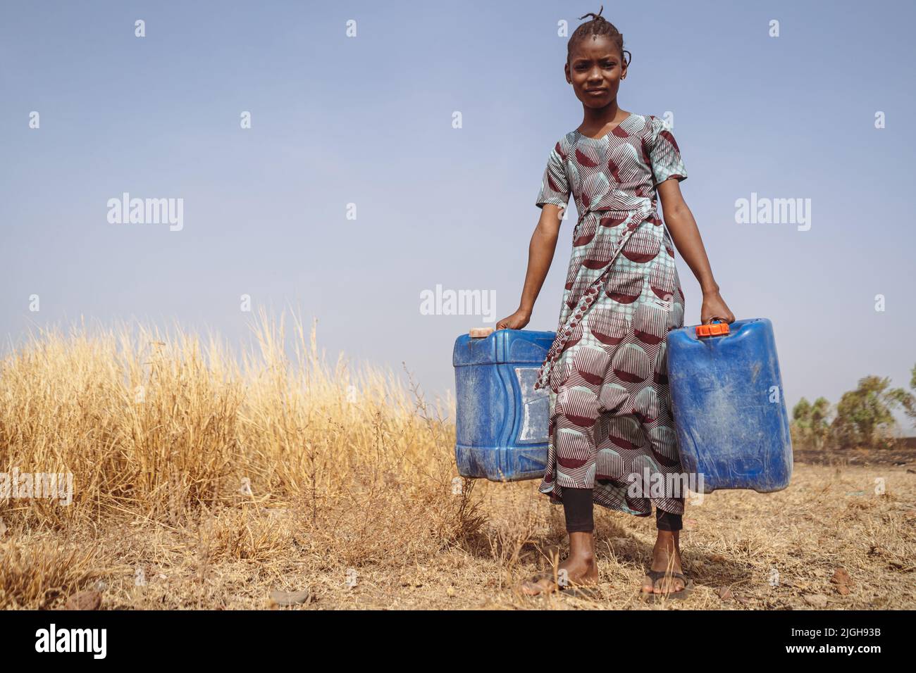 African teenager with two blue water cans walking through a field of dried up grass to get water from the public pump; global warming concept Stock Photo