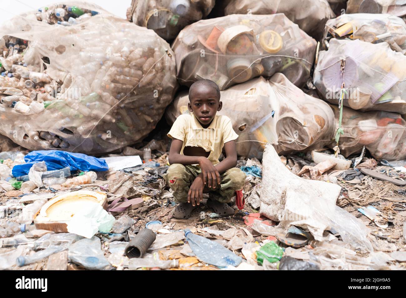 Young Africanboy sitting in front of huge bags full of sorted recyclable plastic bottles at a garbage dump; rudimentary waste management in developing Stock Photo