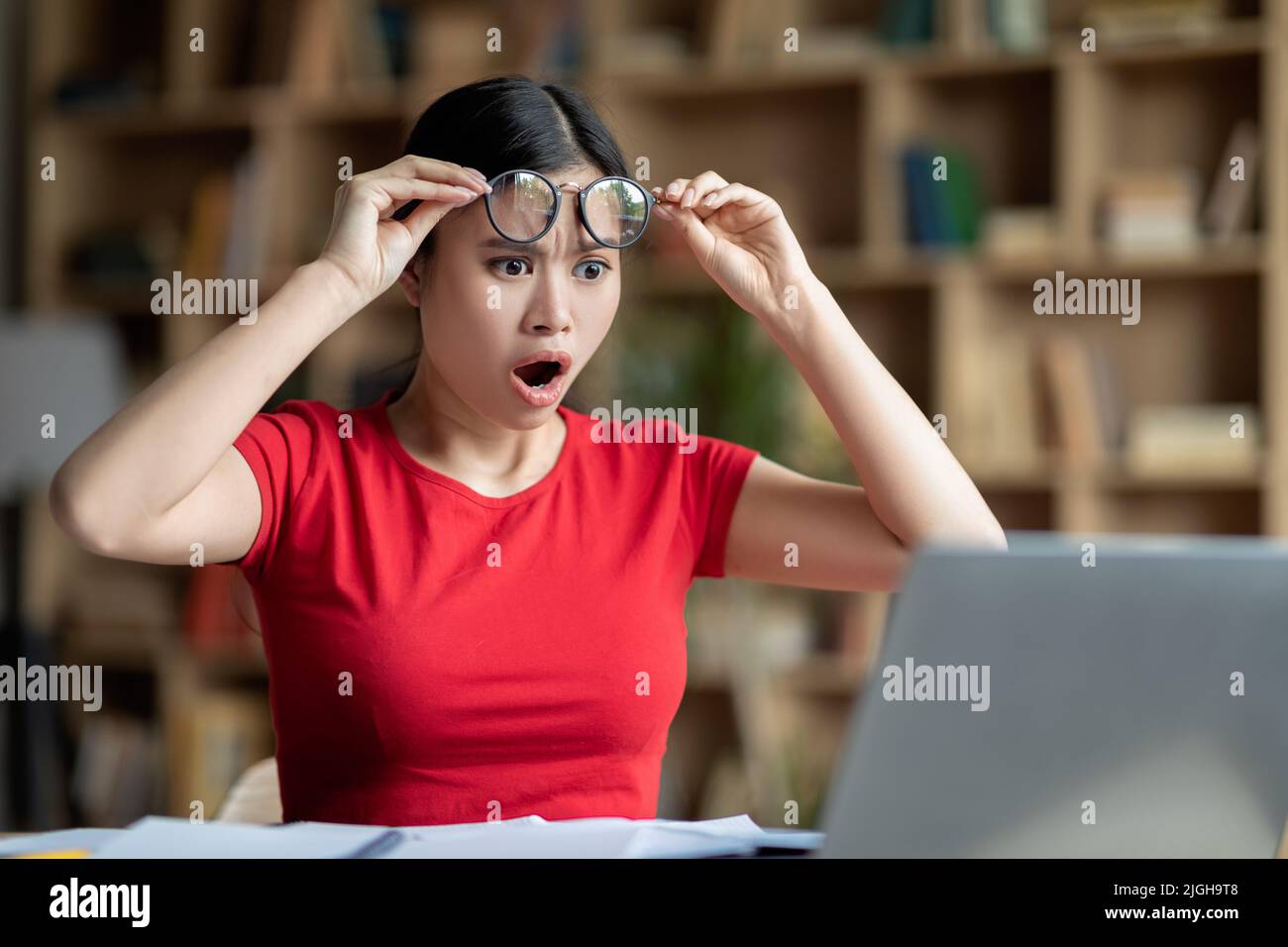 Sad shocked adolescent chinese girl with open mouth takes off glasses looks at computer in room interior Stock Photo