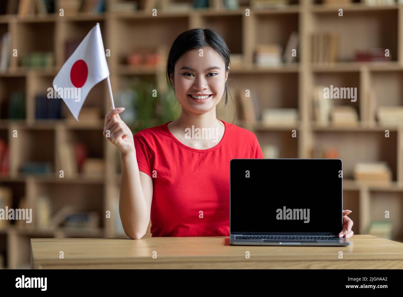 Happy adolescent asian lady student hold Japan flag at table with computer with blank screen in room interior Stock Photo