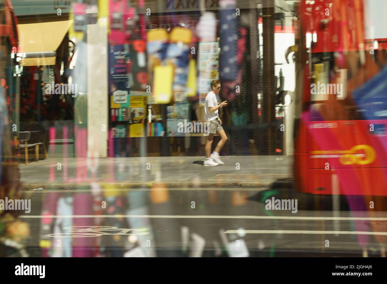 A man walks down new Oxford Street looking at his phone. Taken through umbrella shop window. Reflections.artistic. London bus.Panning Stock Photo