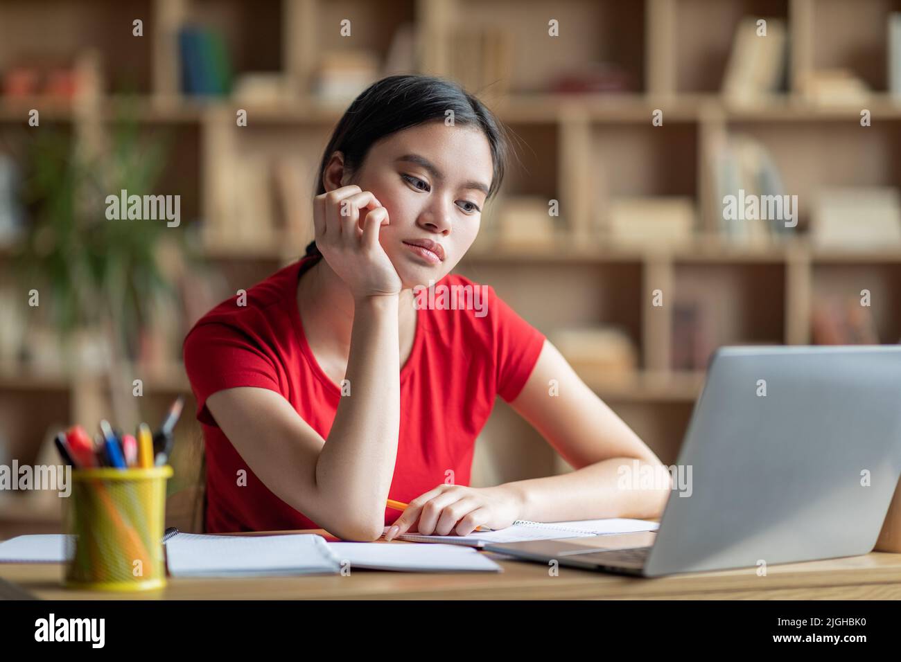 Boring sad adolescent asian female student watching video lesson, lecture on computer in room interior Stock Photo
