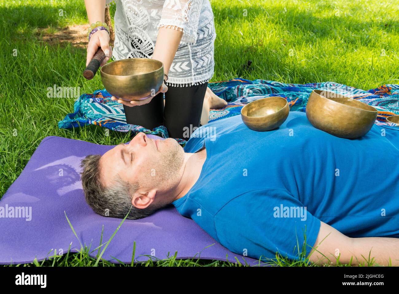 A man lying on a yoga mat with singing bowls on his body while a woman performs a music therapy session with singing bowls on a grassy area. Stock Photo