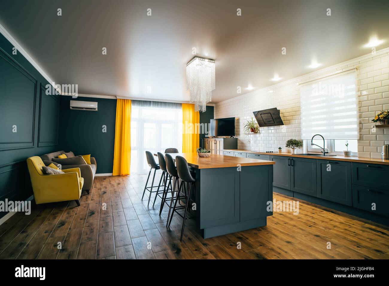 Interior of modern kitchen in blue with yellow accents and wooden floor with large table in middle of room, cooking area and sofa for relaxing. Stock Photo