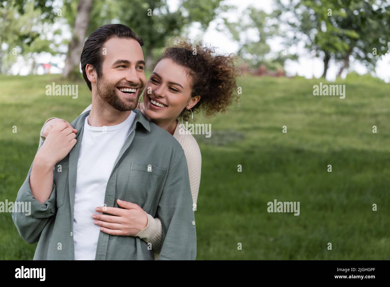 pleased and curly woman hugging boyfriend from behind in green summer ...