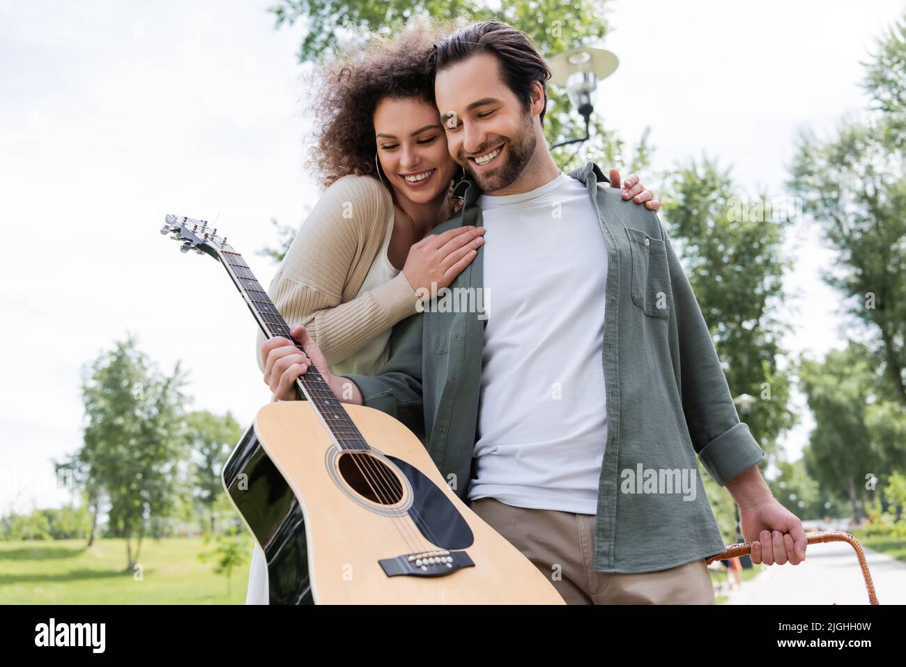 smiling man in summer clothes holding guitar near happy girlfriend in green park Stock Photo