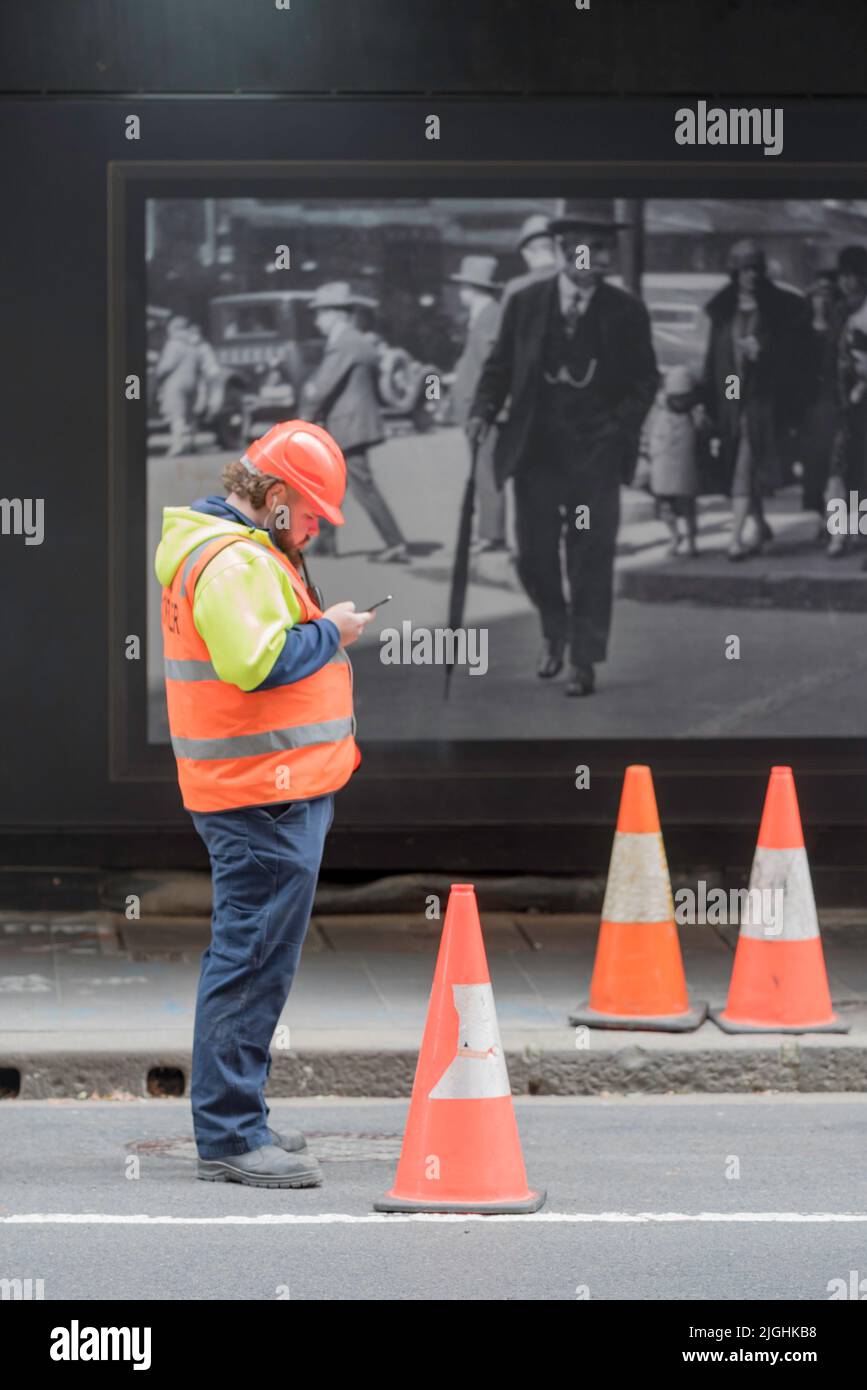 A man stands in Sydney wearing high vis clothing and a hard hat, looking at his phone. In the background an old photo of a man in a suit and hat Stock Photo