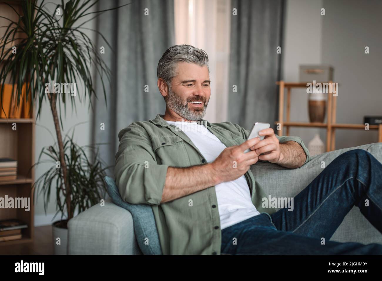 Smiling caucasian mature man with beard watching video, playing game on smartphone Stock Photo