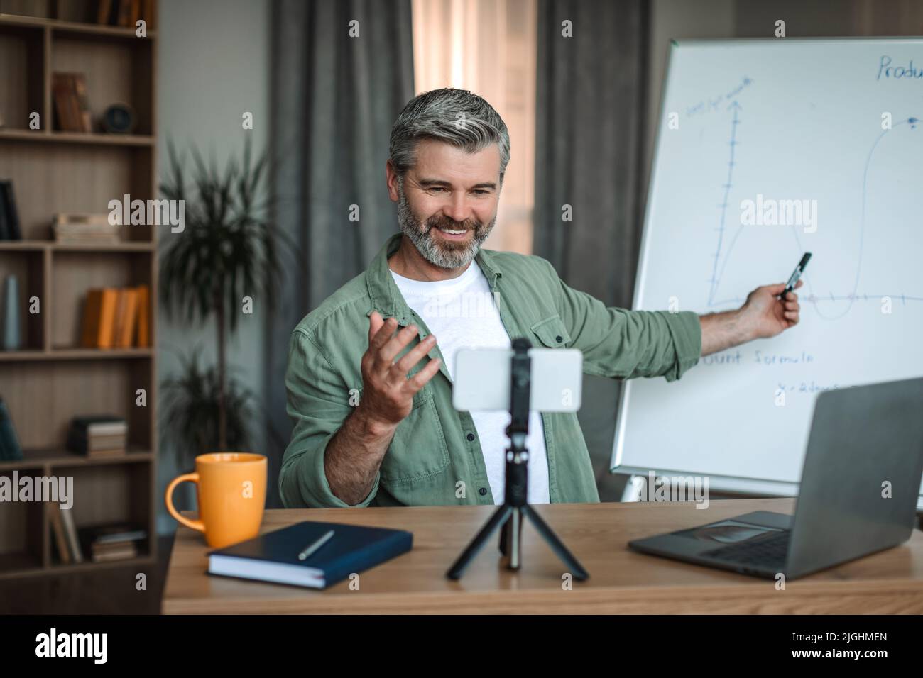 Smiling caucasian old man teacher with beard looks at smartphone and points at blackboard Stock Photo