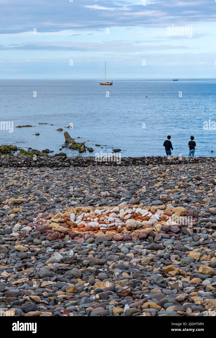 Stone or rock land art pattern or artwork on beach with yacht at sea and boys skimming stones, Berwickshire, Scotland, UK Stock Photo