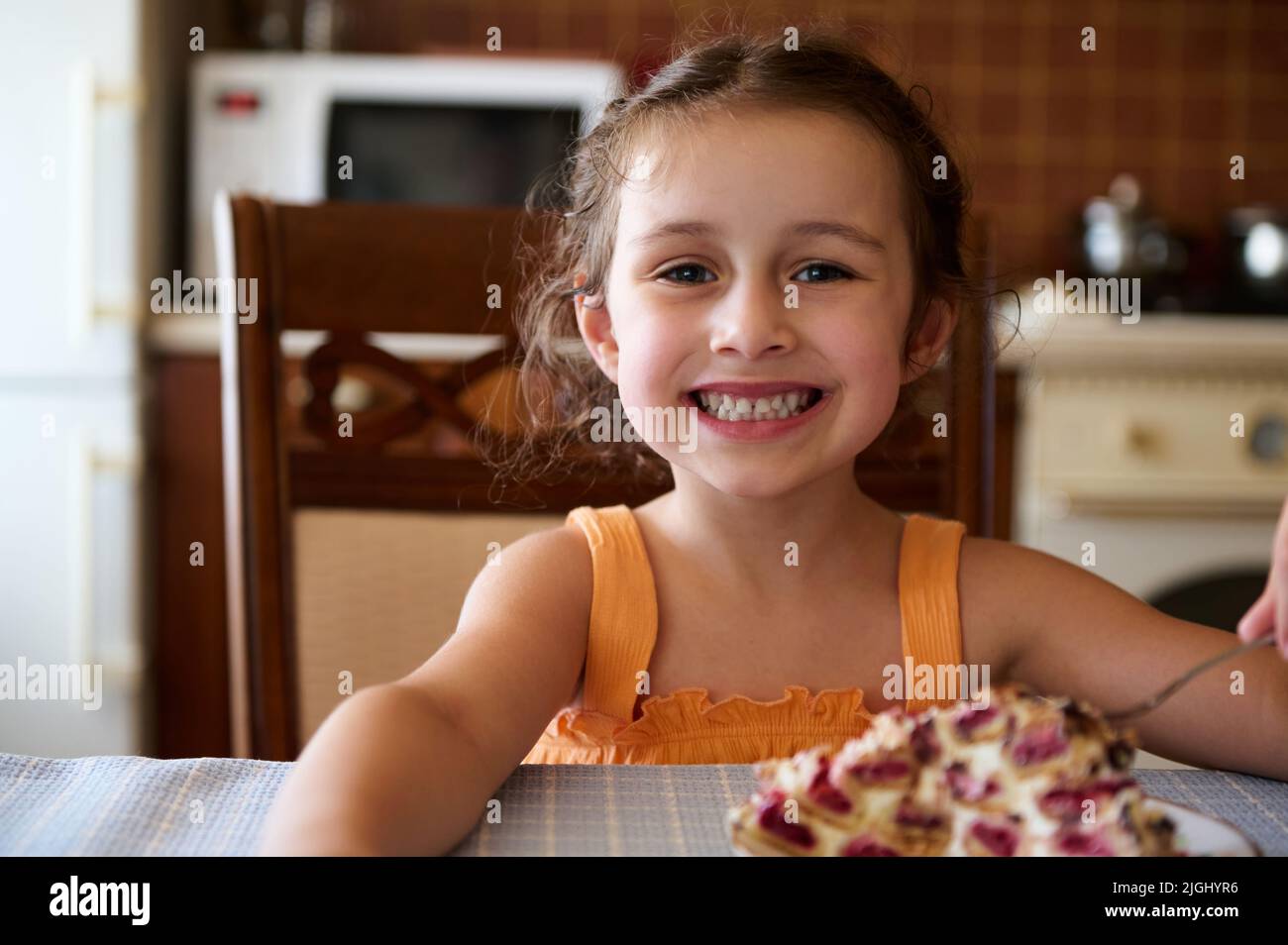 Caucasian little girl smiles, looking at camera, eats a pie from baked dough filled with fresh cherries - Monastery hut Stock Photo