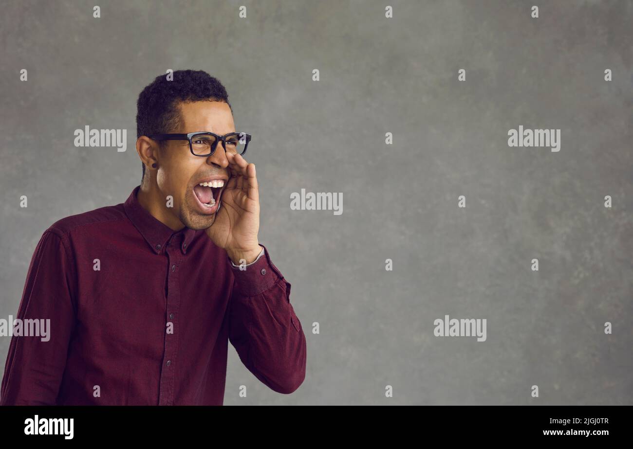 African american man stands on a gray background and shouts holding his hand to his mouth. Stock Photo