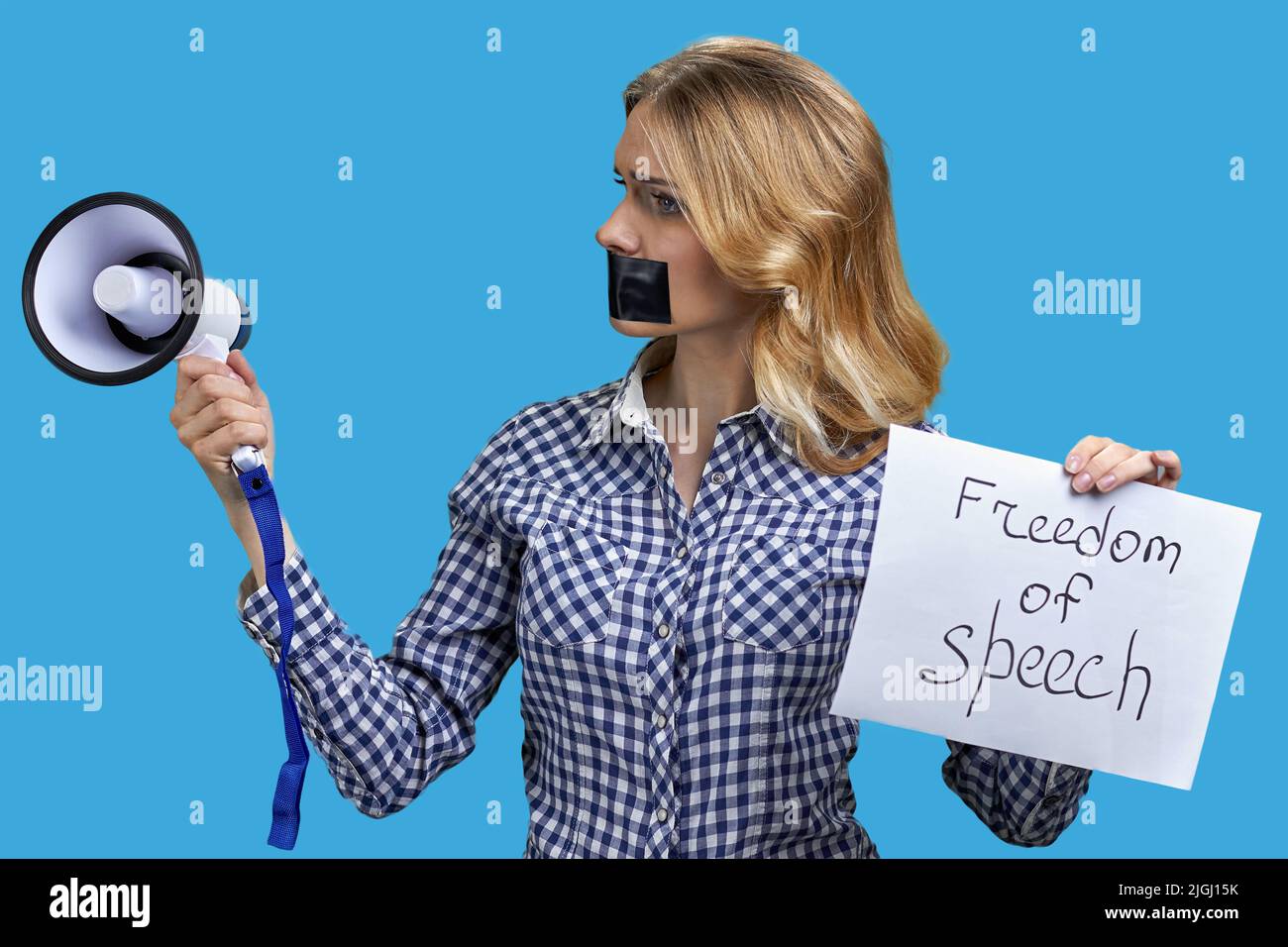 Young speechless woman with adhesive tape on mouth unable to speak in megaphone standing on color background. Freedom of speech. Stock Photo