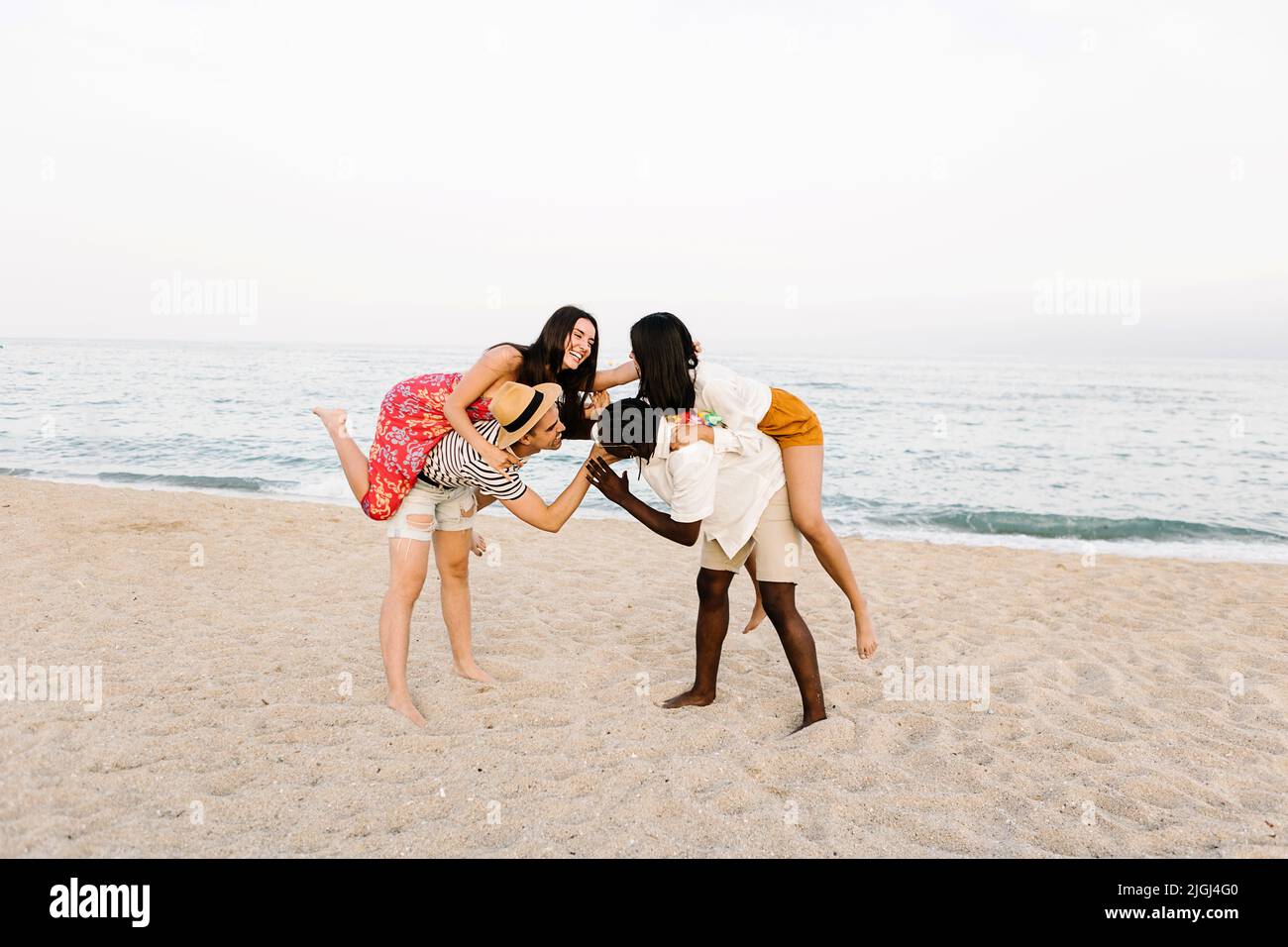 Two multiracial men fighting while piggybacking their girlfriends at beach Stock Photo