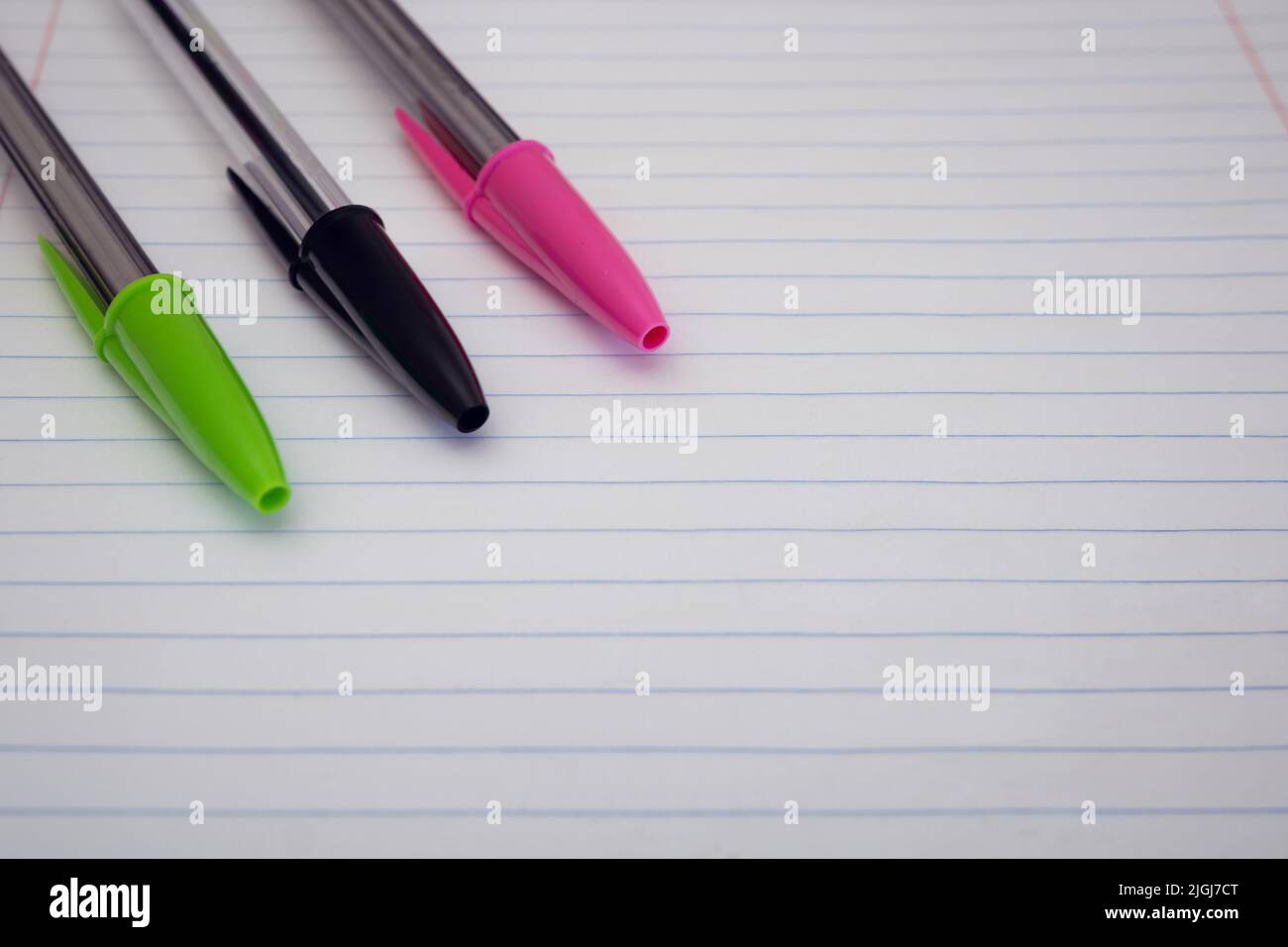Close-up of three pens with caps on resting on a lined paper sheet. Background of green, black and pink pens with selective focus and empty space Stock Photo