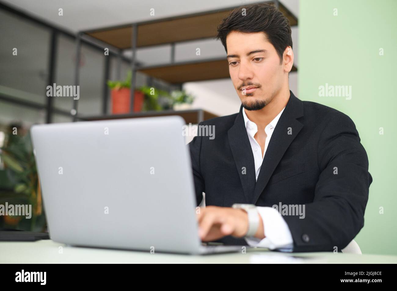 Serious concentrated determined young bearded office worker in business casual clothes, sitting at the desk, typing on laptop, businessman professional is working on project, writing email to client Stock Photo