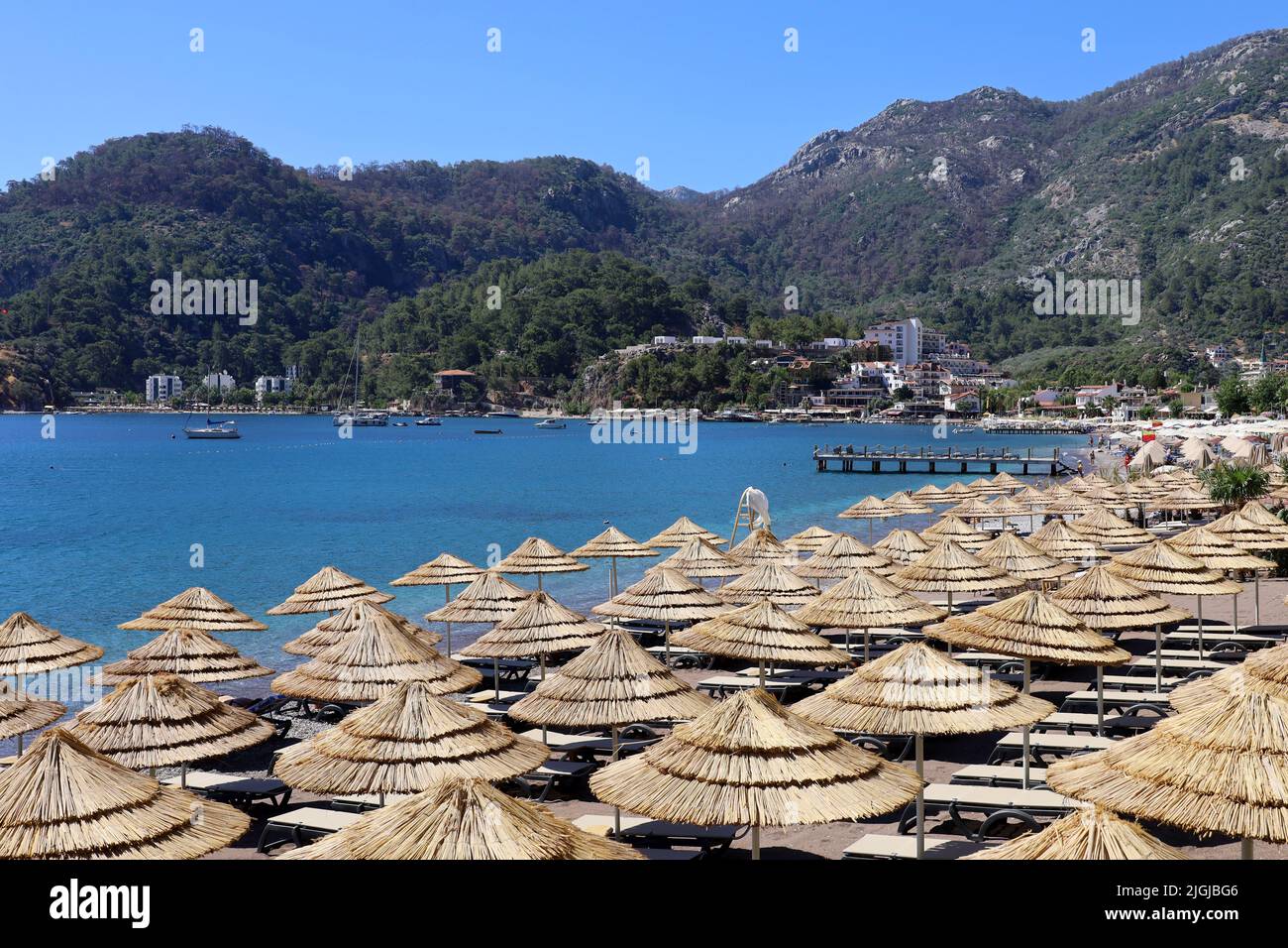View to azure bay and beach hotels surrounded by green mountains. Summer resort with wicker parasols and lounge chairs in Mediterranean sea Stock Photo