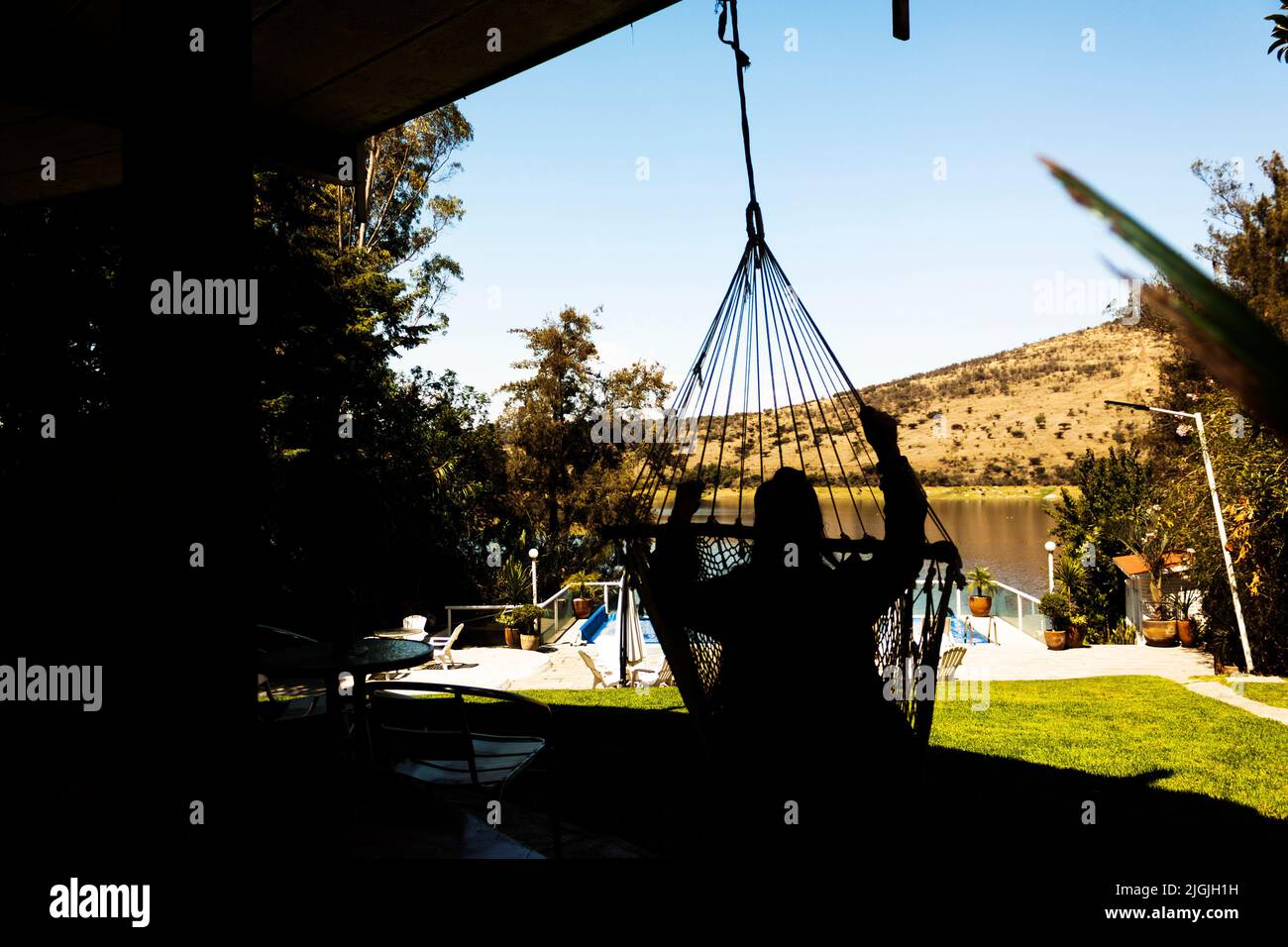 In shadow silhouette of woman sitting on the woven chair of a swing looking towards the lake Stock Photo