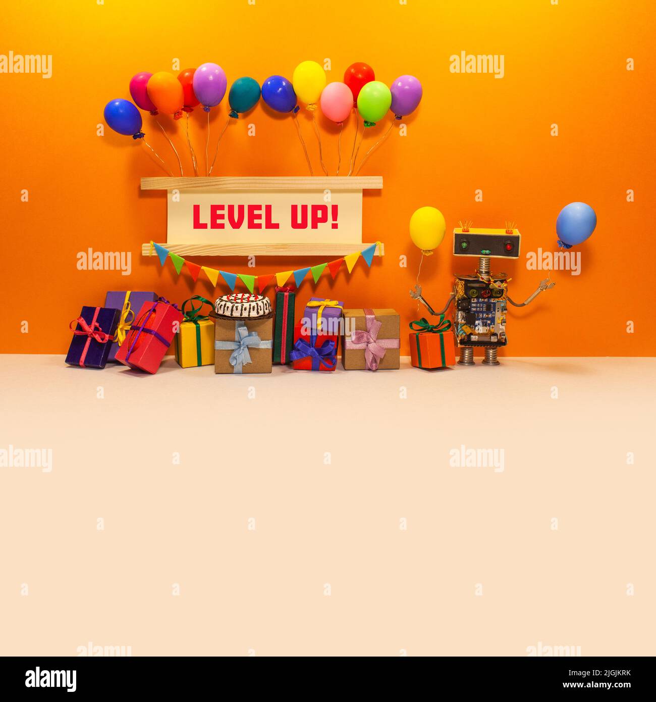 The concept of a holiday birthday card, a toy robot with balloons and gifts near a sign decorated with flags and colorful balloons. Message: Level Up. Stock Photo
