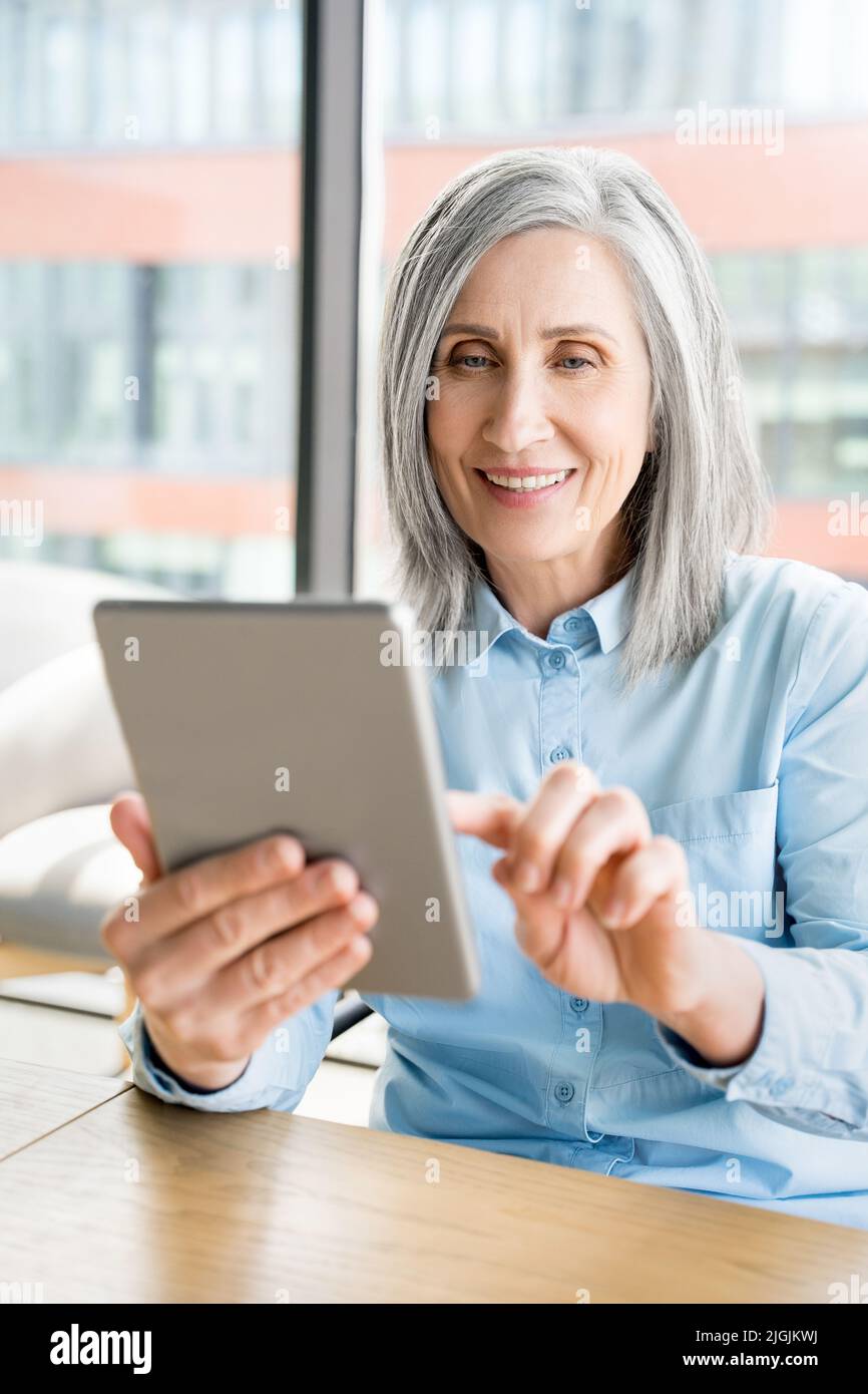 Vertical portrait of old mature woman sit in office and use digital tablet Stock Photo
