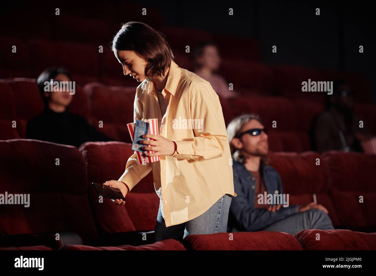 Young woman with popcorn and mobile phone looking for her seat in