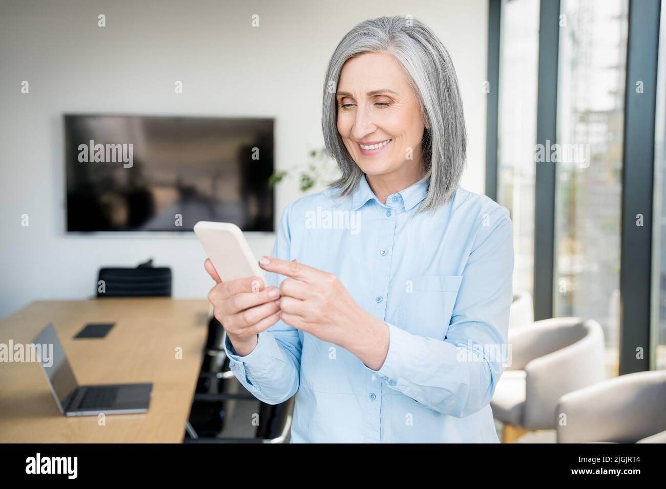 Gray-haired senior businesswoman is using a smartphone in an office. Stock Photo